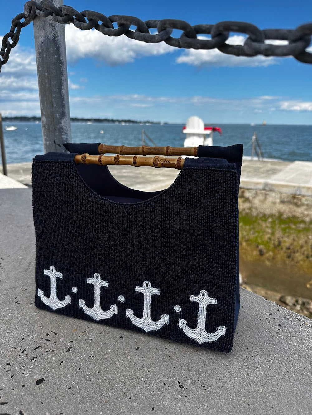 Navy Purse with White anchor boarder, bamboo handle, sitting on cement with ocean behind it. 