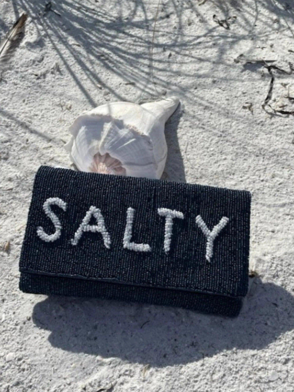Navy beaded clutch with "Salty" in white lettering.  Clutch sitting agains a shell on the beach
