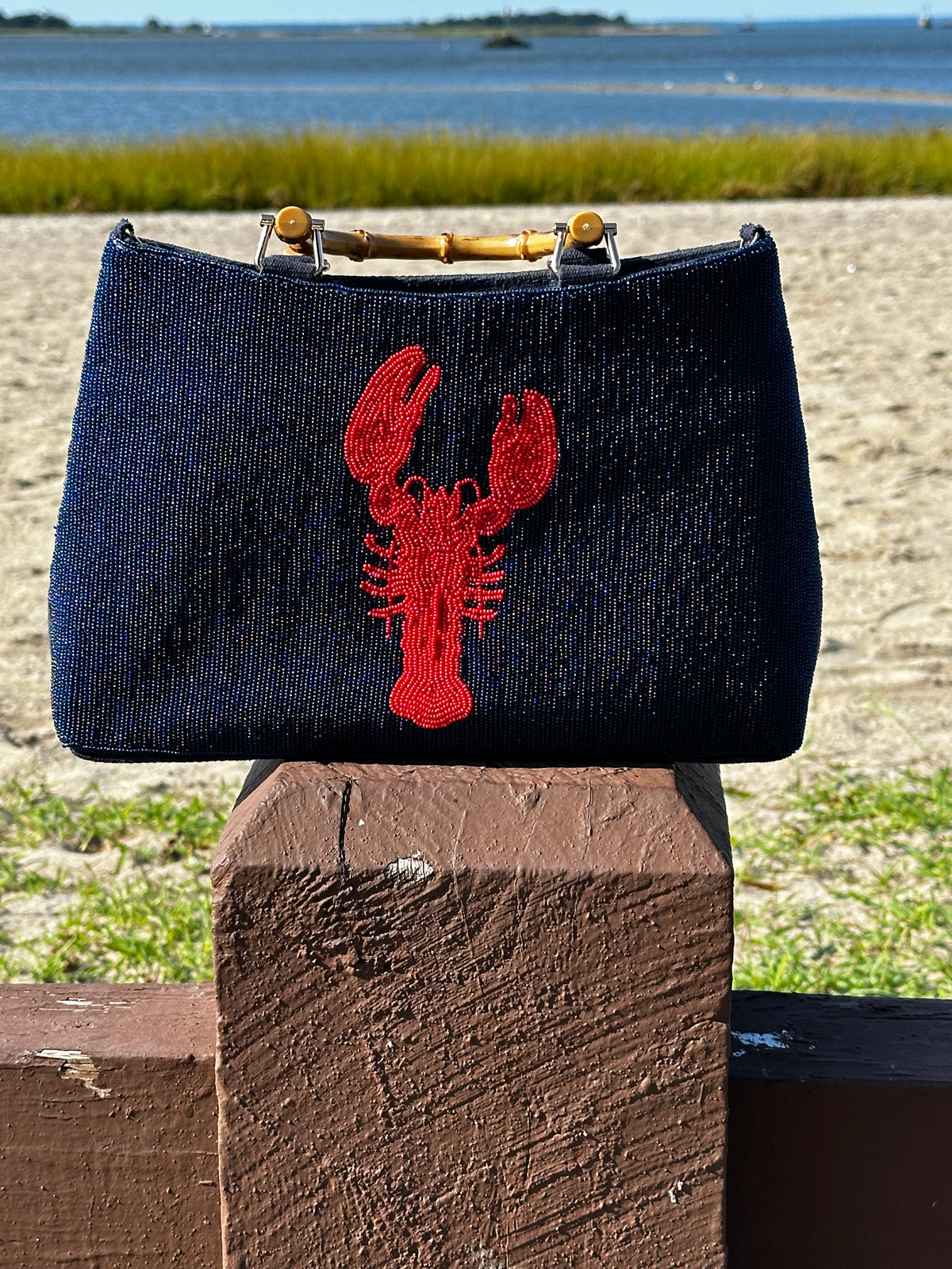 A navy beaded purse with a white lobster design displayed on a bamboo handle, placed on a wooden surface with a beach and foliage in the background.
