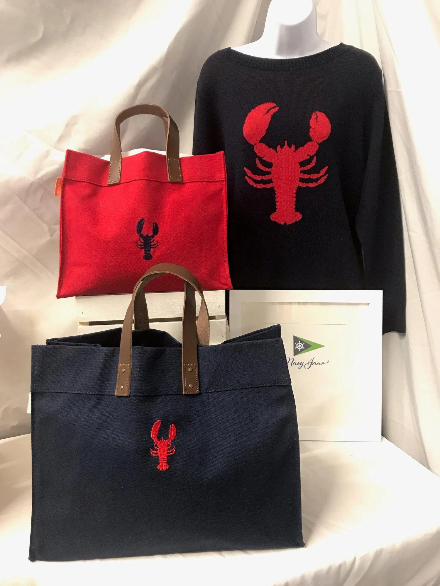 Picture of navy lobster sweater with red lobster.  Also shown is large navy lobster canvas bag with red lobster. Small red canvas bag with navy lobster. 