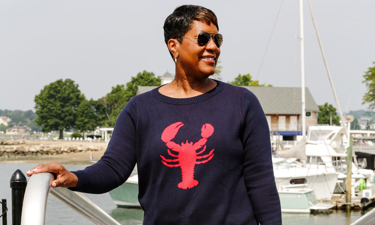 Picture of black women wearing preppy navy sweater with lobster on it. Women is walking up ramp at a dock.  Water is behind her. Sizes are inclusive S-3XL   