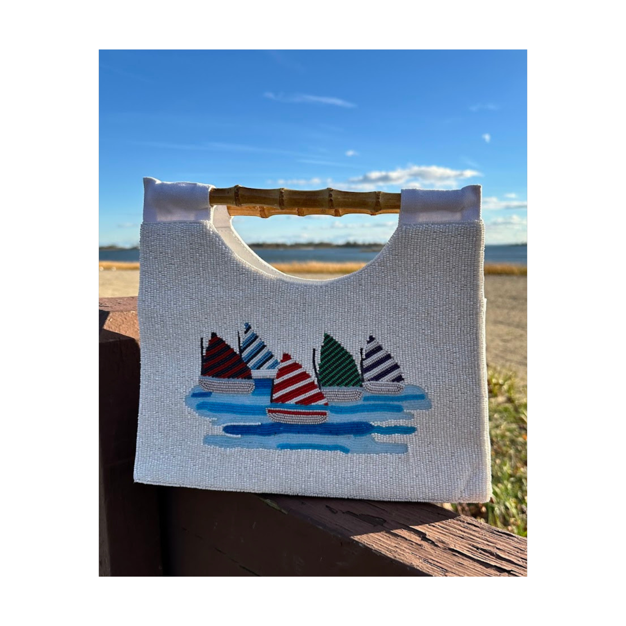 White  beaded bag with sailboat design and bamboo handle against a scenic background