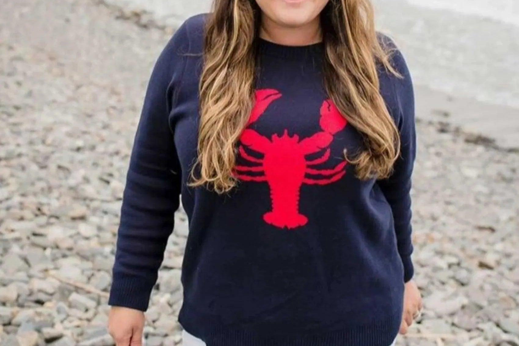 close up of Woman wearing a navy sweater with a red lobster design on a rocky beach.