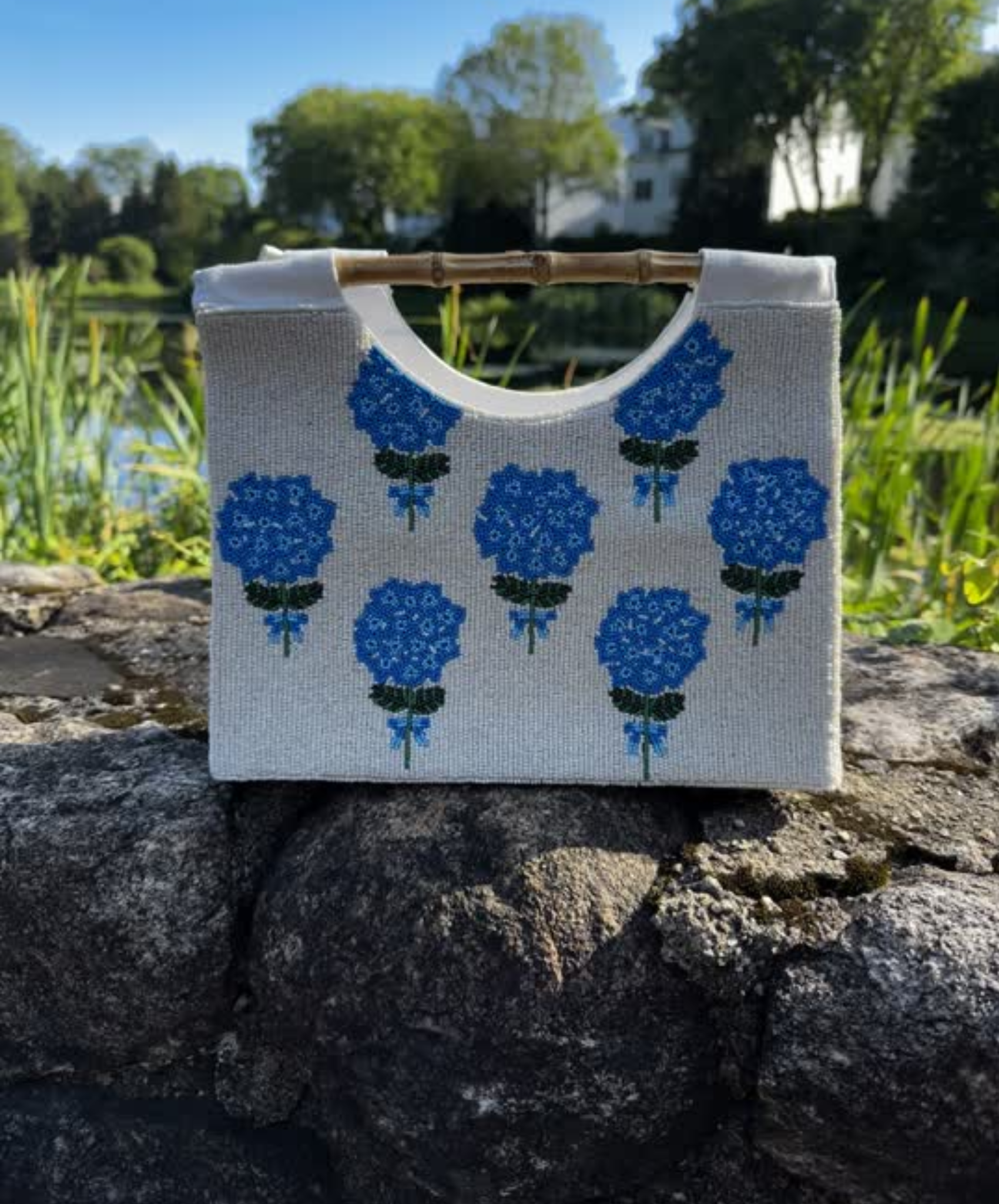 Ceam and blue hydrangea  beaded handbag with straight bamboo handle.  sitting on top of a rock and with grass . 
